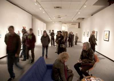 People viewing artworks at an art gallery exhibition. Some visitors are walking and others are seated, with blurred motion suggesting activity. Artworks hang on well-lit walls.