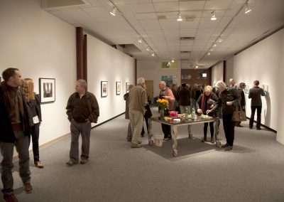 An art gallery event where visitors are viewing photographs displayed on walls while some converse in groups, with a refreshment table in the center.