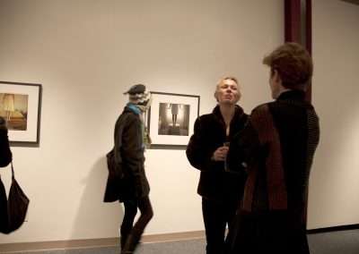 Two people engaged in conversation in an art gallery, surrounded by photographs displayed on the wall, with a third person observing the art.