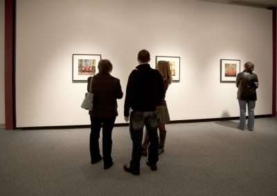 Visitors viewing framed photographs in an art gallery, with each person standing and observing different artworks on white walls under soft lighting.