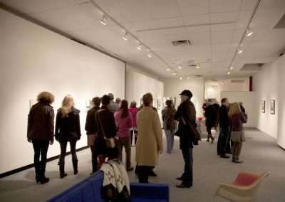 Visitors viewing artwork at a gallery exhibition, wandering and discussing art pieces displayed on white walls under soft lighting.