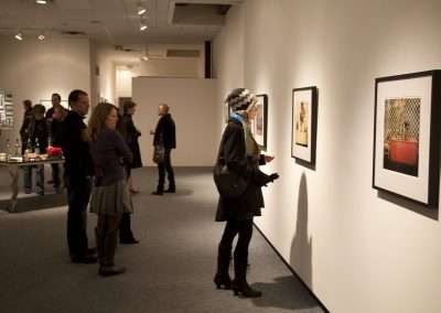 People viewing artwork at an exhibition in a gallery with well-lit framed photographs on the walls. Some attendees are standing, while others are examining the art closely.