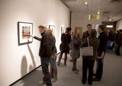People viewing and discussing artwork in a gallery, with focus on one individual pointing at a framed photograph on the wall.