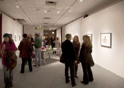 People gather and converse at an art gallery opening, with artwork displayed on the walls of a well-lit corridor. There is a refreshment table in the background.