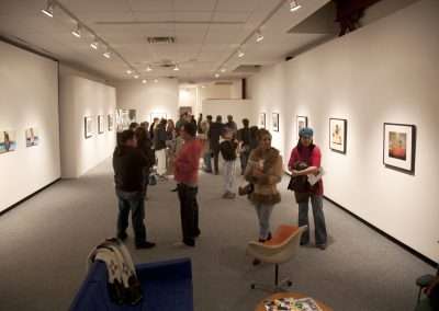 Visitors viewing various framed artworks in a well-lit gallery with white walls and a central seating area with modern chairs.
