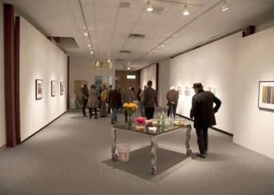 Visitors viewing artworks at an indoor art gallery exhibition; some are walking while others stand and observe, with a table of refreshments in the foreground.