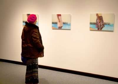 A person in winter attire views three framed photographs of a figure by the beach, displayed on a white wall in an art gallery.