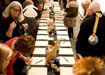 People gather at a busy art gallery auction, examining various small artworks displayed on a table, while holding drinks and engaging in conversation.