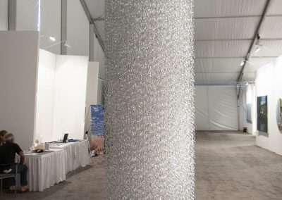 A large, glittering column wrapped in shiny silver sequins stands in the center of a tented art exhibition hall, with booths and artworks visible in the background.