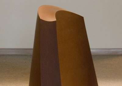 A minimalist sculpture by Tom Waldron of a curved, rusty metal sheet forming a loop, displayed on a light carpeted floor against a plain wall with focused lighting.