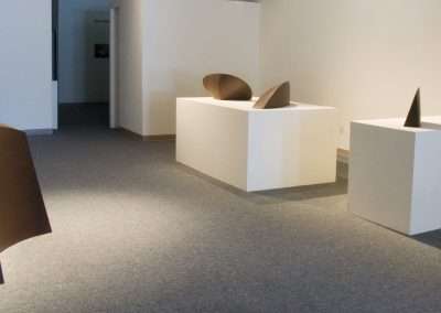 Modern art exhibit featuring minimalist sculptures by Tom Waldron, resembling curved metal shapes on white pedestals in a gallery with carpeted floors and white walls.