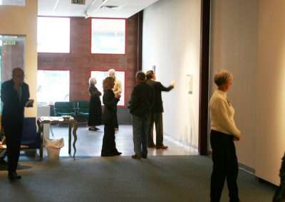 People gathered in a brightly lit hallway, interacting and looking at items displayed on the walls. Some individuals stand alone, while others engage in conversation.