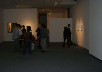 People mingling and viewing art at an indoor gallery exhibition with refreshments on a table in the foreground and artworks displayed on white walls.
