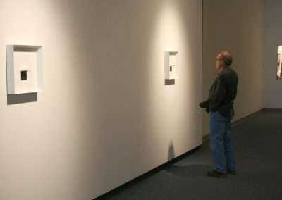 A man observing abstract art pieces in a minimalist gallery with white walls and focused lighting. The art is composed of square frames within frames.