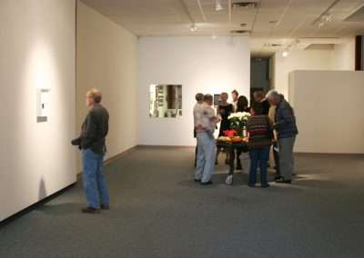 People viewing artwork in a modern gallery setting with white walls. A group gathers near a table with flowers, while others observe pieces individually.