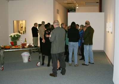 People mingling at an indoor art gallery event, with artwork displayed on walls and a refreshments table to one side.