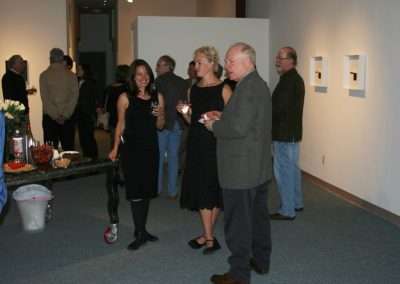 People socializing at an indoor art gallery opening. A group is casually conversing with drinks in hand near a table with refreshments, surrounded by artworks displayed on the walls.