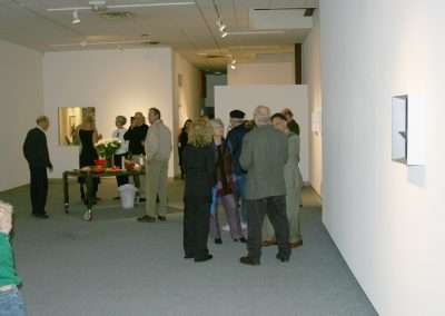People viewing artwork at a gallery opening, mingling and discussing the exhibits, with bright lighting and sparse modern decor.