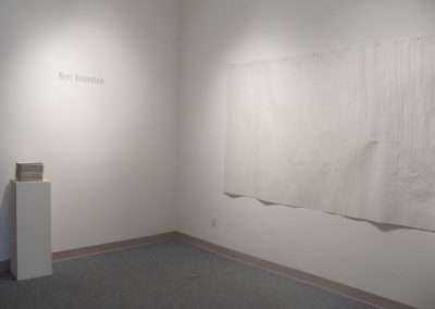 An art gallery installation featuring a plain, textured white canvas hanging on a wall, a stack of papers on a pedestal, and an artist's name, Kerri Rosenstein, displayed on the opposite wall.