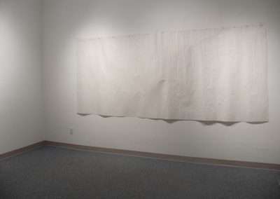 A large white textured canvas hangs on a light gray wall in a sparsely furnished room with a speckled gray carpet. The canvas is unevenly rectangular and looks slightly crumpled.