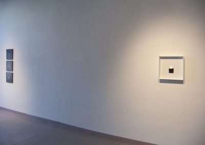 Modern art gallery wall featuring two minimalist art installations, one a series of four small square panels, and the other a single framed piece, under subtle lighting.