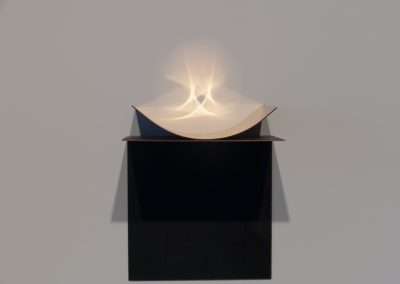 A conceptual art piece featuring a folded paper projecting a radiant light pattern on the wall, displayed on a black pedestal against a light-colored background.
