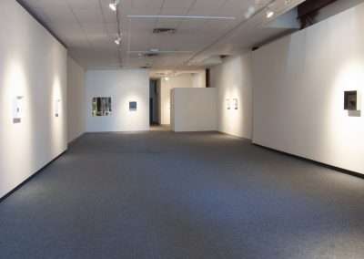 An empty modern art gallery with white walls displaying various framed artworks, and a gray carpeted floor leading through the illuminated interior.