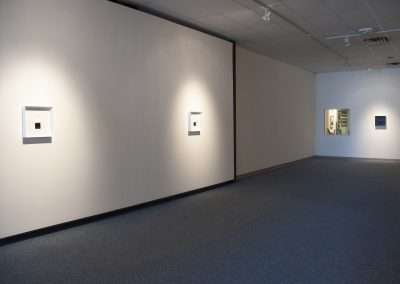 A modern art gallery with white walls displaying small, framed abstract artworks, each illuminated by individual spotlights, in a quiet and spacious interior setting.