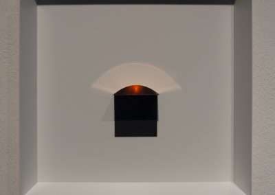 A minimalistic modern art display featuring a white shallow box frame on a wall, containing a black semi-circle with a small rectangular form beneath it, both partially lit by warm lighting.