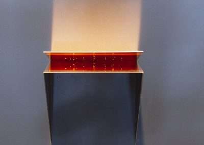 A modern black pedestal with a bright red interior illuminated from within, casting a golden light upwards against a gray background.