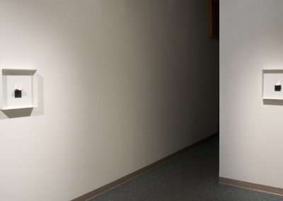 A minimalist gallery hallway featuring two small, framed artworks displayed in wall niches, with soft lighting accentuating the pieces.