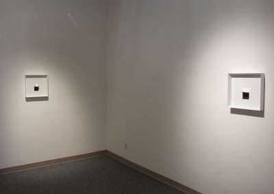 An image of a minimalist art gallery with two small square artworks centered on separate walls in a neutral-toned room with soft lighting.