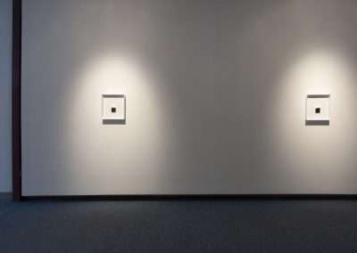 A minimalist gallery wall with two illuminated square frames showcasing small white art pieces, spaced evenly against a soft gray background.
