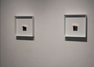 Two white-framed niches on a gray wall, each displaying a small bronze sculpture resembling a shell illuminated from above.