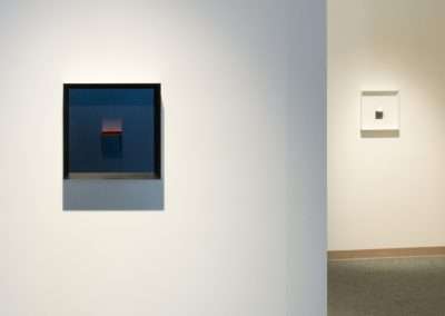 A minimalist art gallery with two framed abstract paintings on plain white walls, one close-up on the left and another at a distance on the right.