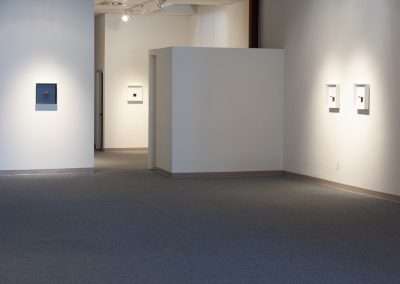 Interior of an art gallery featuring white walls adorned with framed artwork, partition walls creating separate gallery spaces, and a well-lit carpeted floor.