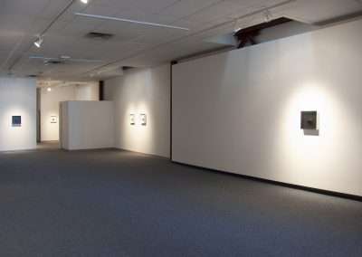 An art gallery with white walls showcasing framed artworks, each illuminated by individual lighting. The gallery features a carpeted floor and an open, spacious layout.