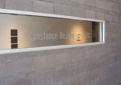 A frosted glass window embedded in a brick wall displays the text "Constance DeJong arc" with an interior view showing additional framed squares through the window.