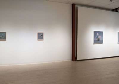 A modern art gallery interior displaying four framed artworks on white walls, each artwork spaced evenly, under soft gallery lighting with polished wooden floors.