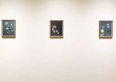 Three framed floral paintings displayed in a row on a soft beige wall in an art gallery. Each painting features different flower arrangements with dark backgrounds.