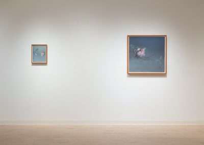 Two framed paintings of flowers hanging on a clean, well-lit gallery wall, one large on the right and one smaller on the left, over a light wooden floor.