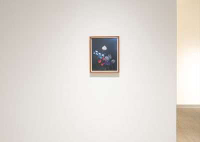A small, framed floral painting hung on a plain, white wall in a brightly lit gallery space. The artwork stands as a focal point in the minimally decorated room.
