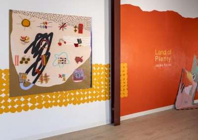 Art exhibition featuring a colorful textile artwork on a wall beside a section painted orange with the title "Land of Plenty" by Jackie Riccio.