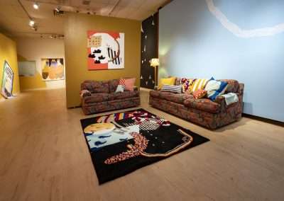 A vibrant art gallery interior featuring a patterned sofa covered in colorful pillows, a decorative rug, and various abstract paintings on the walls.