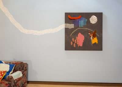 A modern art installation featuring a textured fabric art piece with abstract designs on a wall next to a patterned sofa with colorful cushions. A white undulating line stretches across the blue wall.