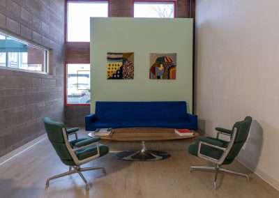 A modern waiting area with a blue sofa, two green armchairs, a wooden oval table, and three abstract paintings on a mint green wall. The room has wooden flooring and large windows.
