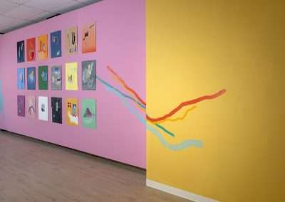 Interior of an art gallery with two walls displaying abstract artwork; one wall is pink with multiple framed pieces, and the other is yellow with a colorful, wavy line painting.