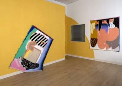 Two large, colorful, abstract art pieces displayed on a yellow wall in a gallery, with one canvas leaning against the wall and another hanging. Light hardwood floors and a window enhance the room.