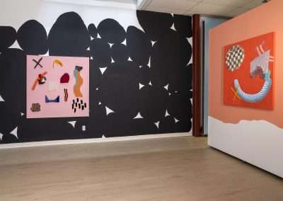 An art gallery room featuring two abstract paintings: on the left, a pink background with assorted shapes; on the right, an orange canvas depicting a surreal scene. A large black mural with white triangles is in the background.