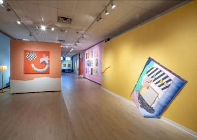 Contemporary art gallery interior featuring brightly colored walls and diverse artwork, including a large angled canvas near the foreground. Soft lighting enhances the vibrant space.